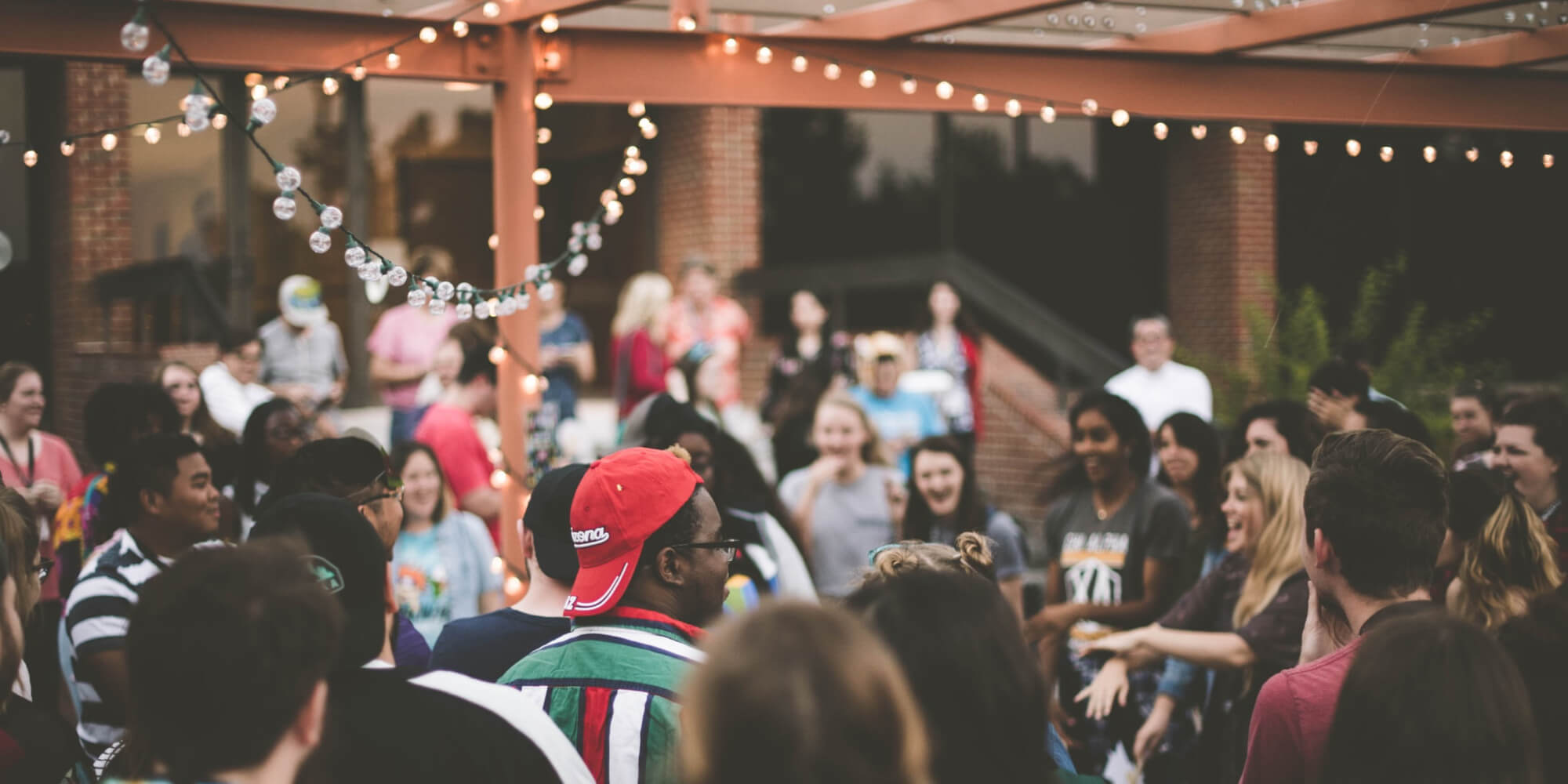 A crowd of people smiling and laughing beneath a covered structure. Strings of lights hang from above.